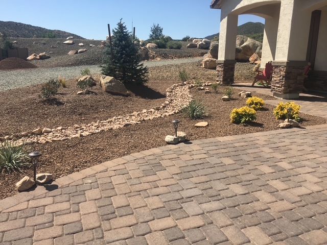 Brick patio and landscaped yard beside a house, with potted flowers and a small tree on a sunny hillside.