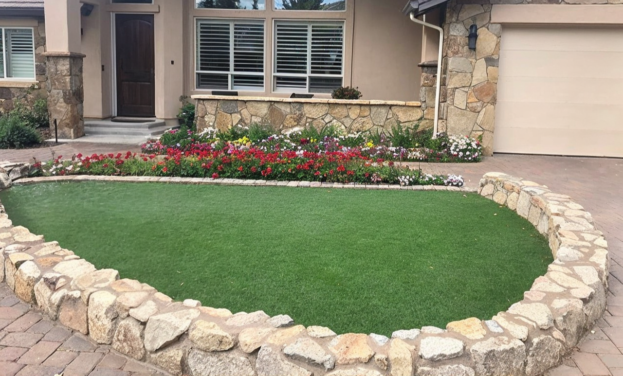 Curved stone-walled garden bed with artificial green turf in front of a house, colorful flowers in background.