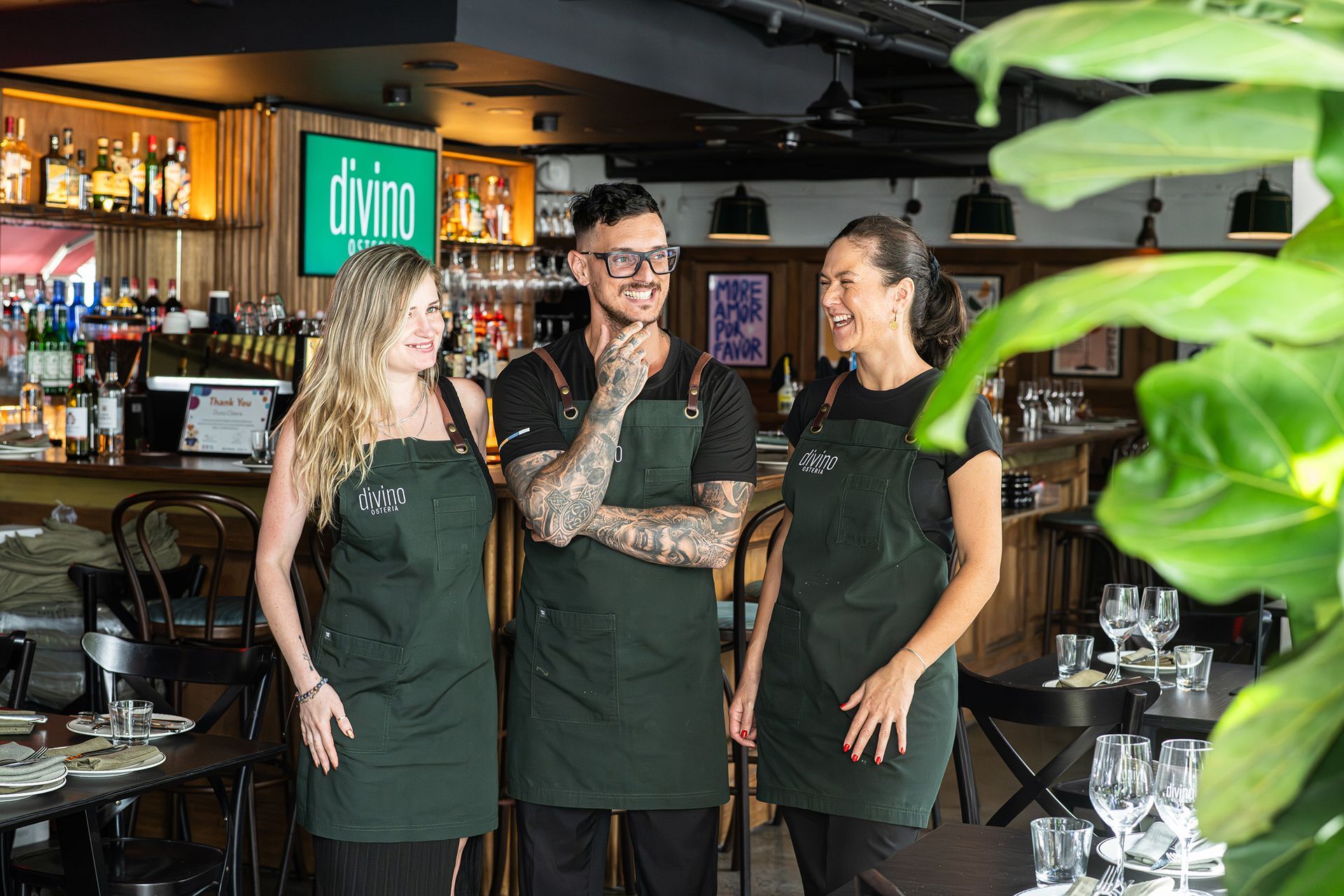 Three restaurant staff smiling, wearing green aprons, in front of a bar with the restaurant name