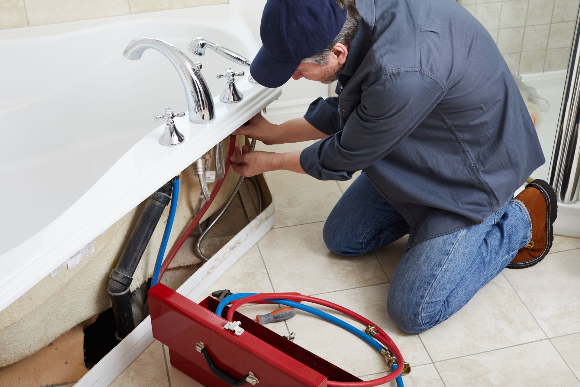 Plumber kneeling under a sink, repairing pipes with a red tool case on the bathroom floor.