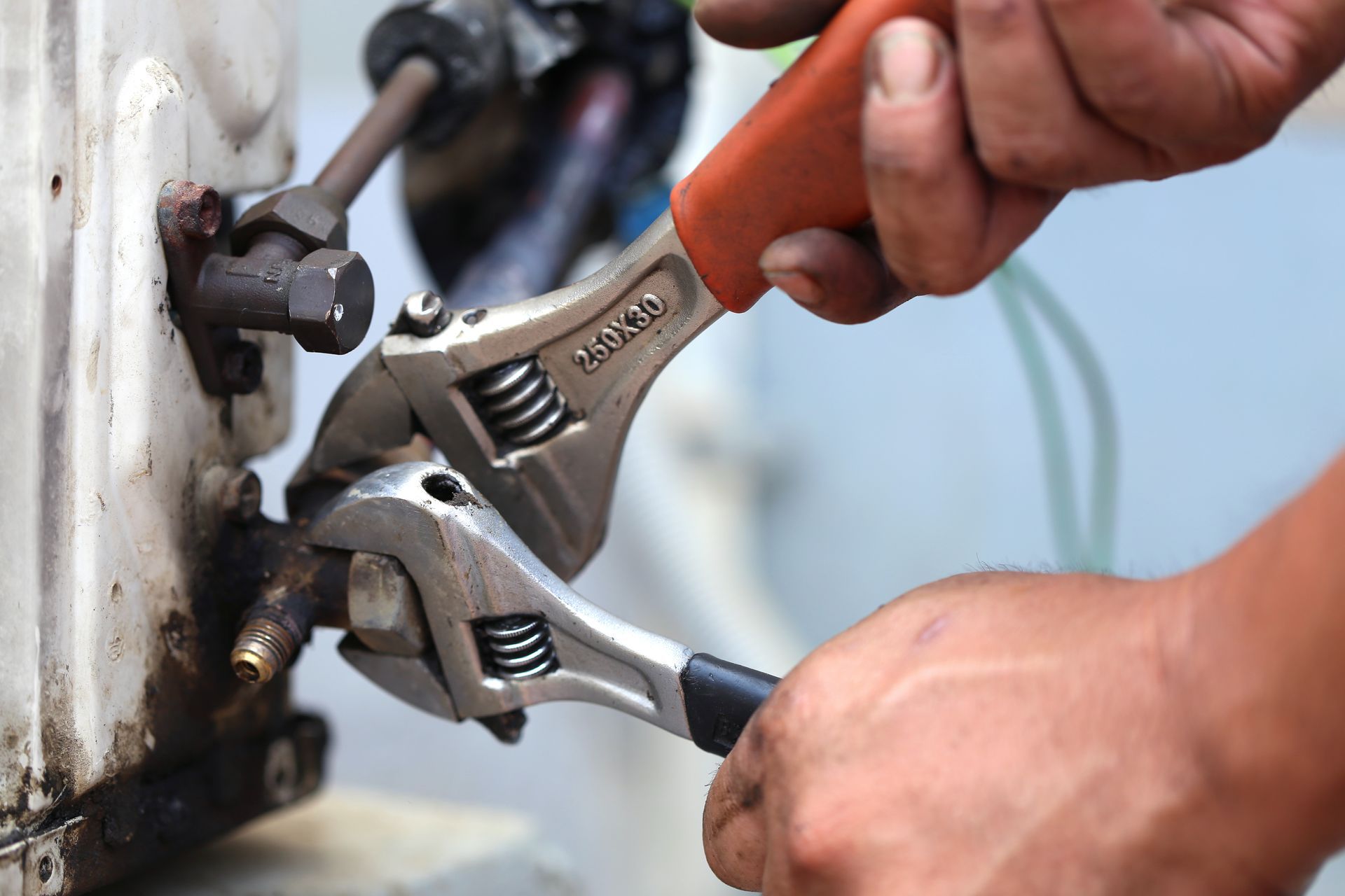 Hands using adjustable wrenches to tighten a metal pipe fitting on a white surface