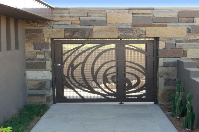 A decorative black metal gate with a swirling design set in a stone wall. A concrete path leads up to the gate.
