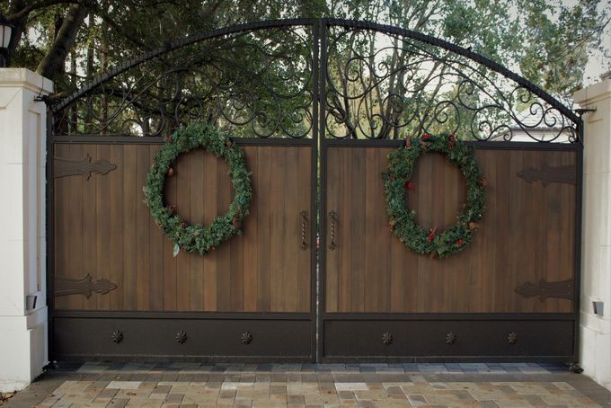 Wooden double gates with arched ironwork, decorated with green wreaths, leading to a brick driveway; a Christmas setting.