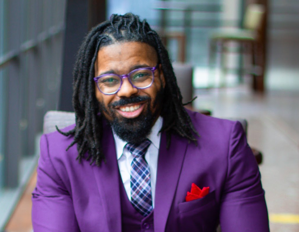 Man with dreadlocks and glasses, wearing a purple suit, smiling in a modern interior.