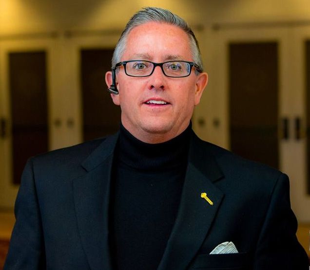 Man wearing glasses and a black turtleneck under a suit jacket, smiling, a gold key pin, indoors.