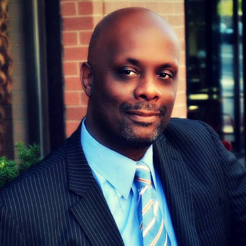 Man in suit and tie, smiling, outdoors near brick wall.