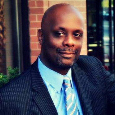Man in suit and tie, smiling, outdoors near brick wall.