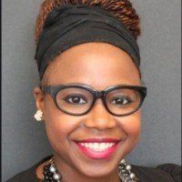Woman with glasses and a black headwrap smiles, wearing a black top and a beaded necklace.