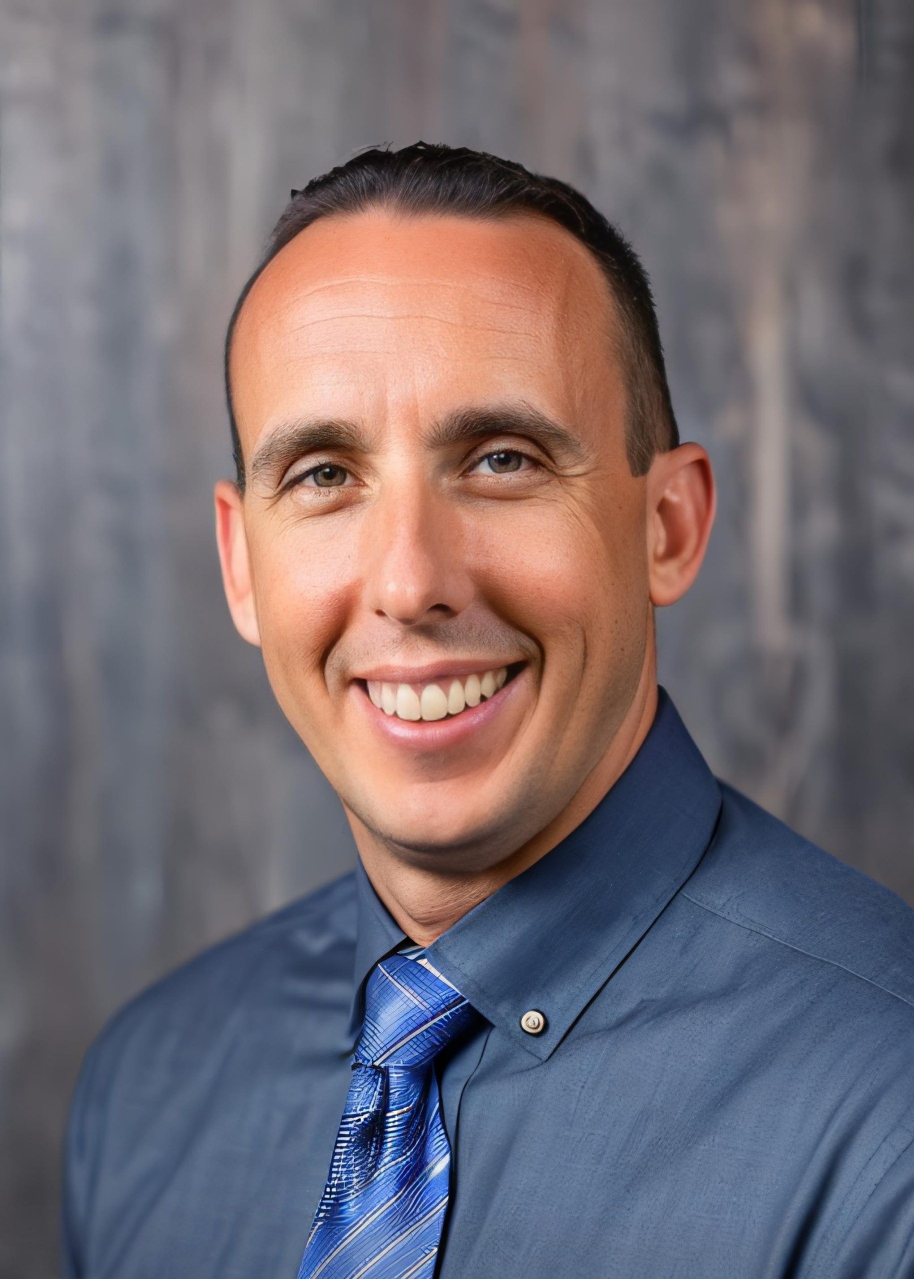Man in blue shirt and tie smiling at the camera against a grey background.