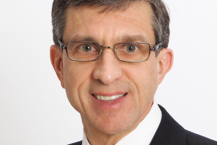 Man wearing glasses, smiling, in a dark suit against a white background.