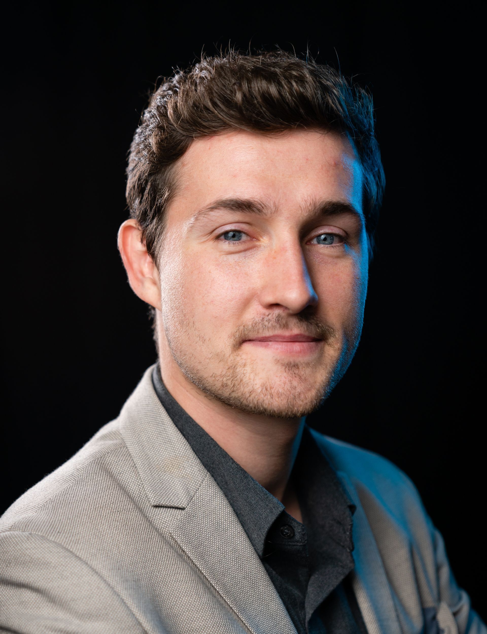 Man in a blazer smiles, looking at the camera against a dark background.