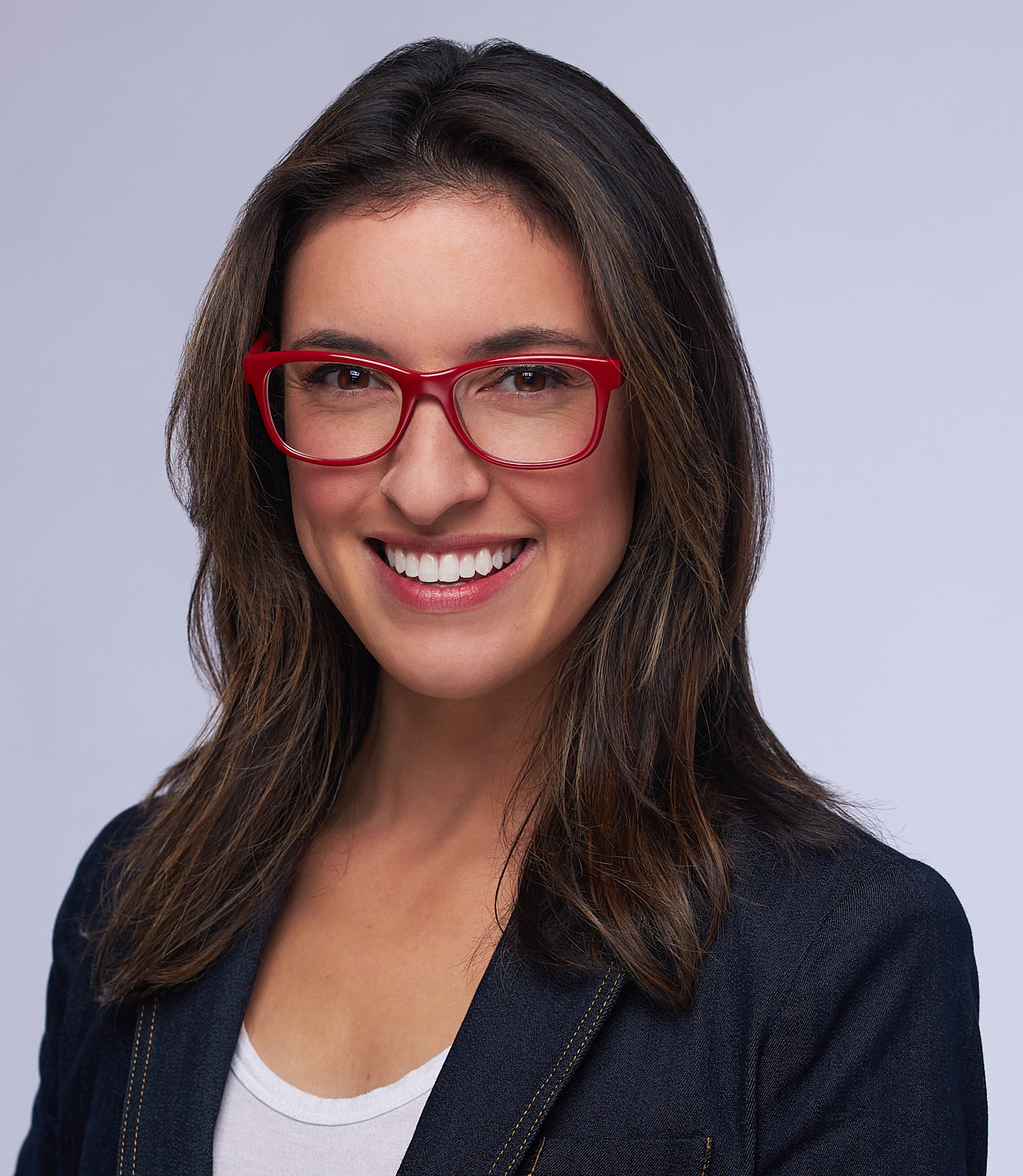 Woman with glasses smiles, wearing a denim blazer, white shirt, and red-rimmed glasses, against a light gray background.