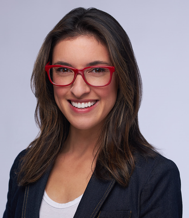 Woman with glasses smiles, wearing a denim blazer, white shirt, and red-rimmed glasses, against a light gray background.