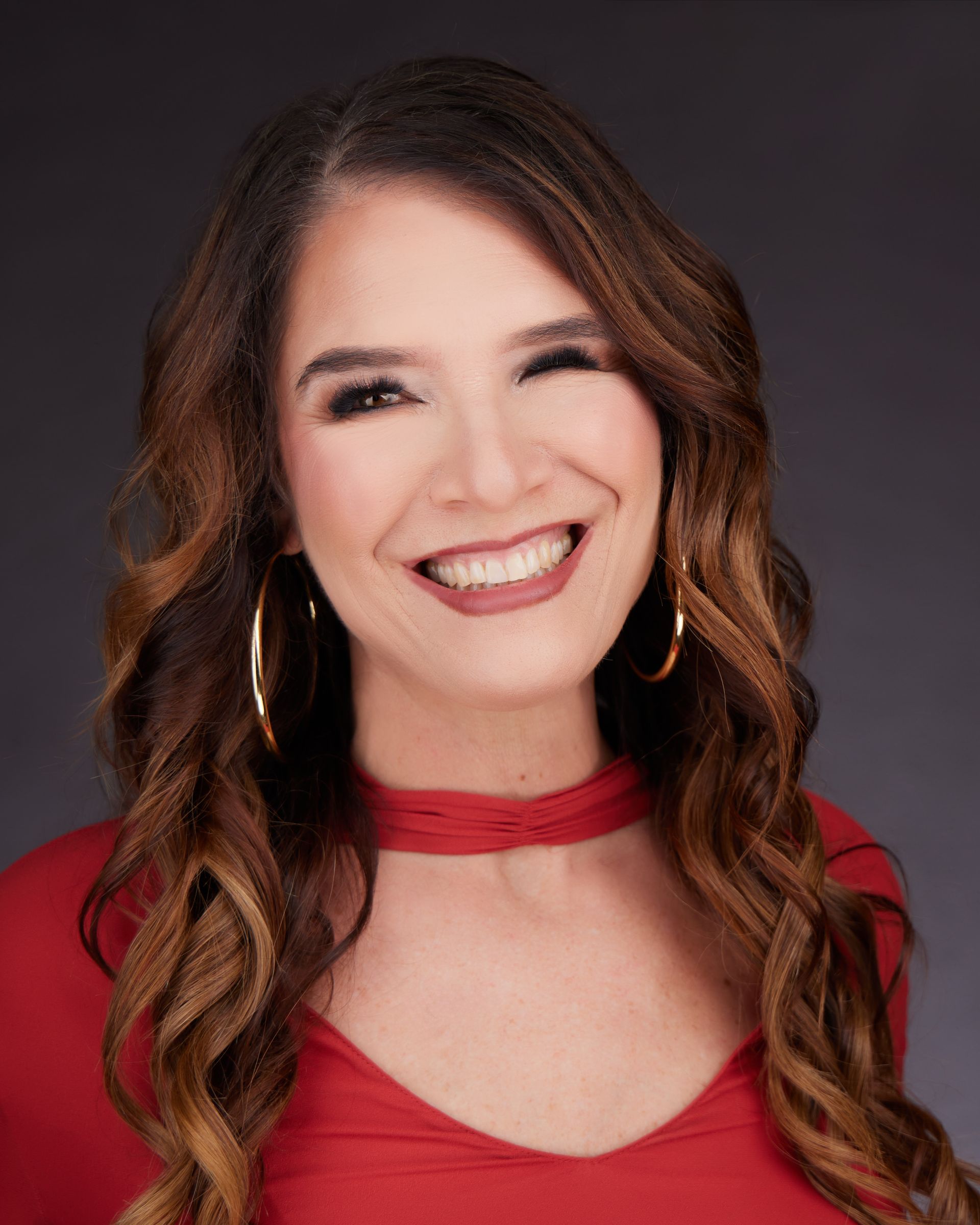 Woman with long brown hair, wearing a red top and large gold hoop earrings, smiles broadly.