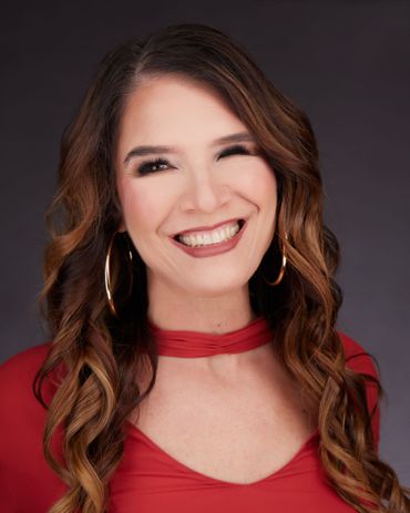 Woman with long brown hair, wearing a red top and large gold hoop earrings, smiles broadly.
