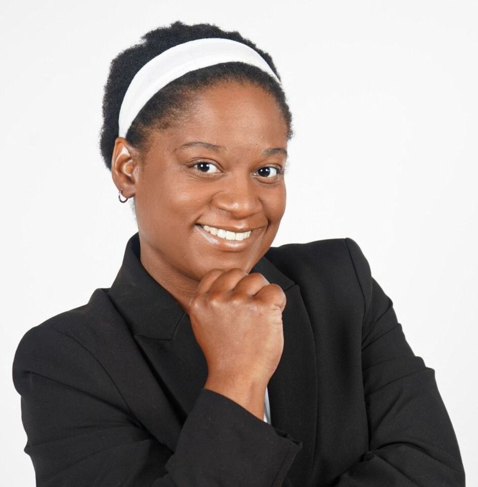 Woman in a black blazer and white headband smiles, resting chin on a fist.