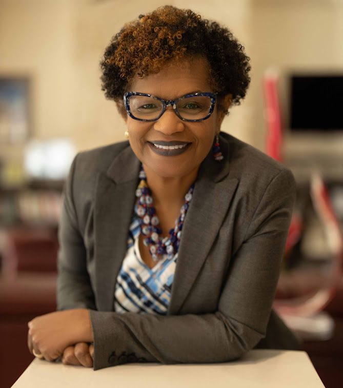 Woman in glasses and gray blazer smiles, leaning on a white surface.