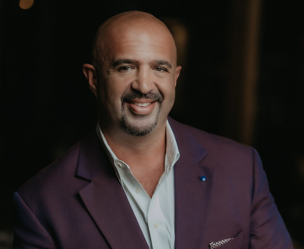 Man in purple blazer smiles at camera, dark background.
