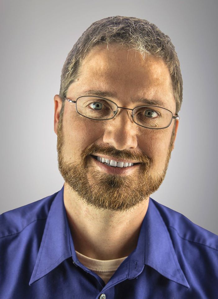 Man with beard and glasses smiles, wearing a blue collared shirt, against a gradient gray background.