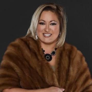 Woman wearing brown fur shawl and black beaded necklace smiles, against a dark background.