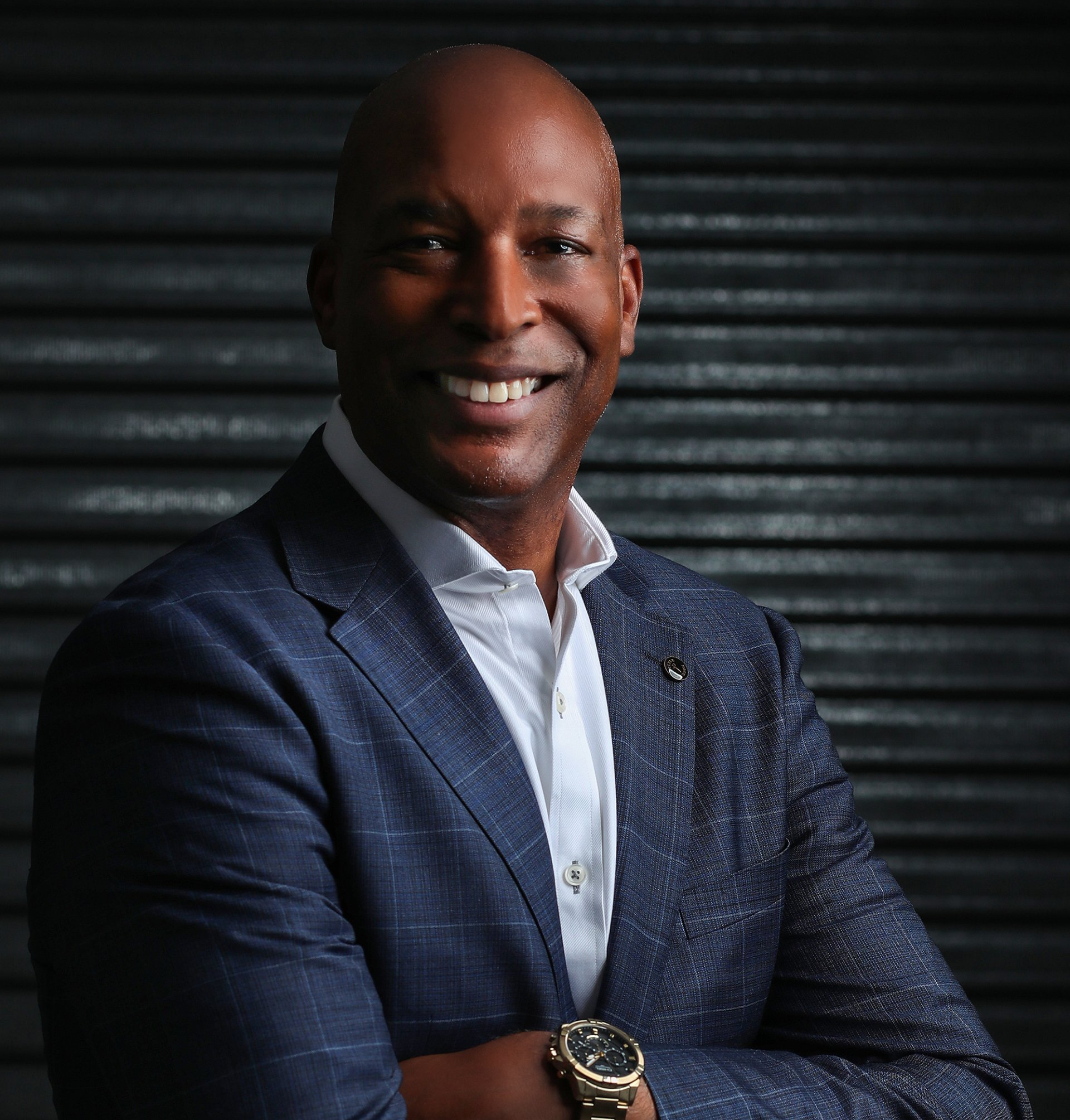 Man in a blue suit smiling, arms crossed, set against a dark textured background. He wears a white shirt and a wristwatch.