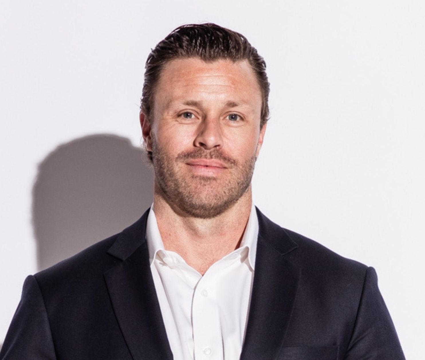 Man in a suit and open-collar white shirt, looking directly at the viewer, set against a plain white background.