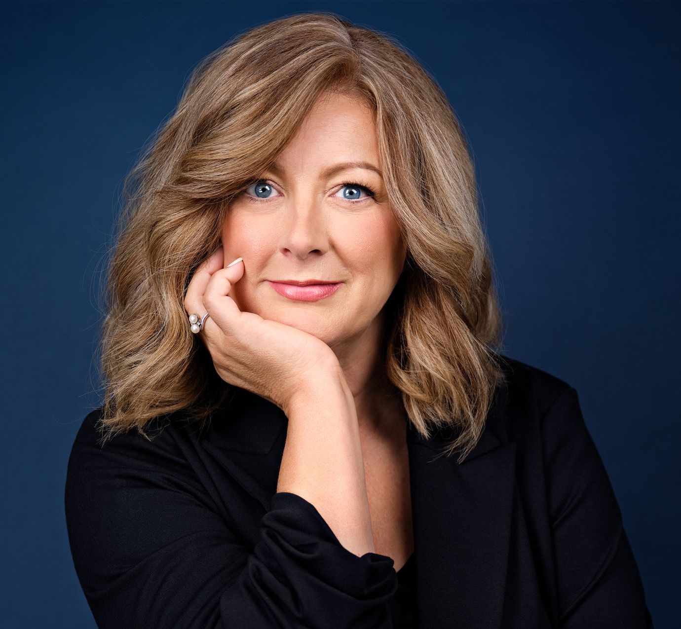 Woman with blonde hair, resting chin on hand, smiling. Dark blazer, blue background.