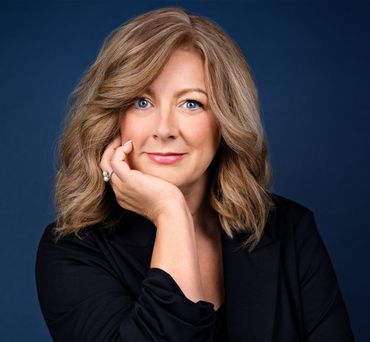 Woman with blonde hair, resting chin on hand, smiling. Dark blazer, blue background.