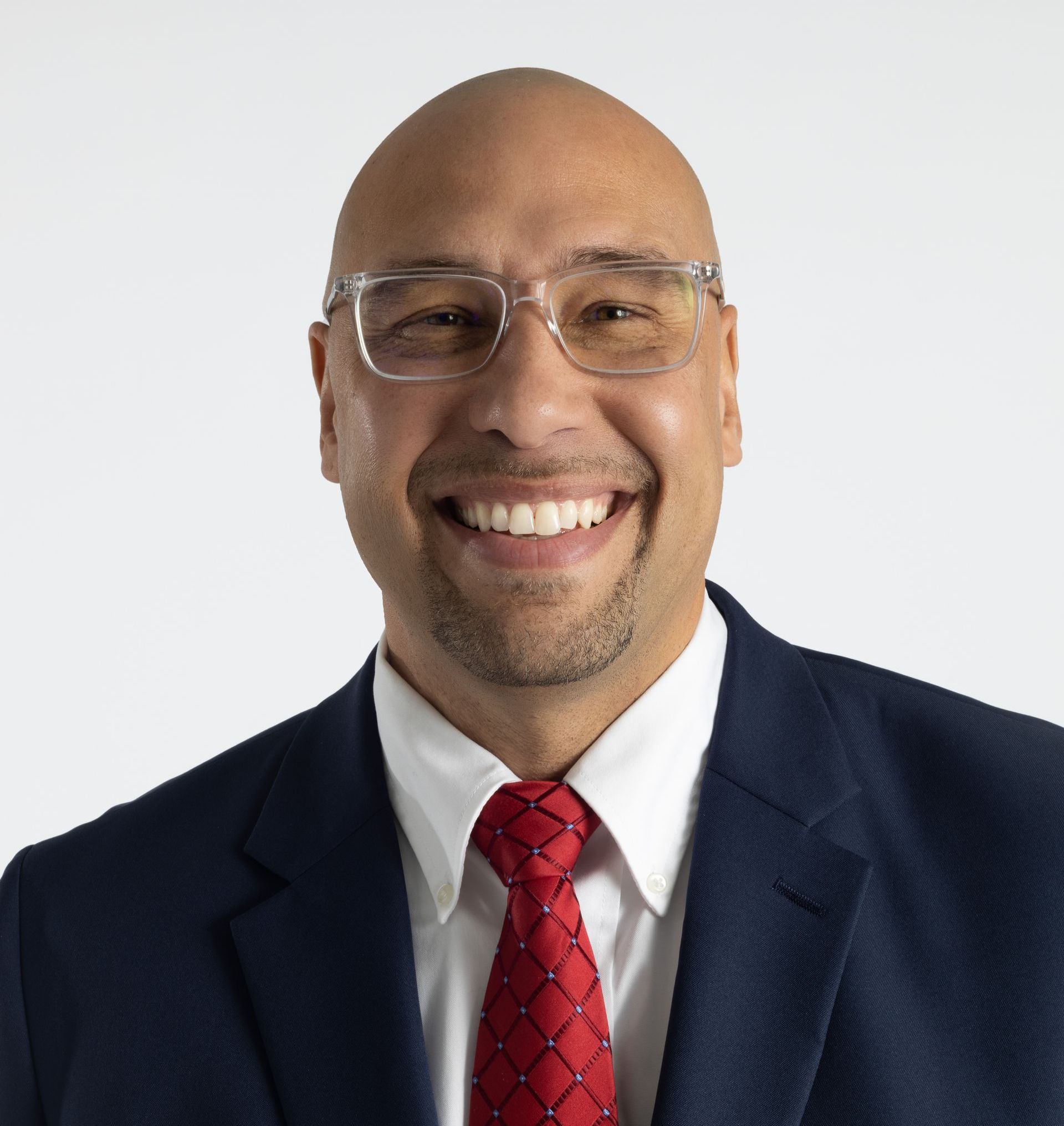 Man with glasses, bald head, and red tie smiling in a navy suit against a white backdrop.