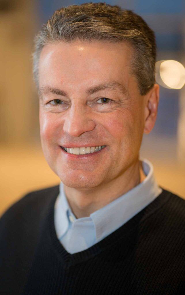 Man smiling, wearing a blue shirt under a black sweater. Blurred background.