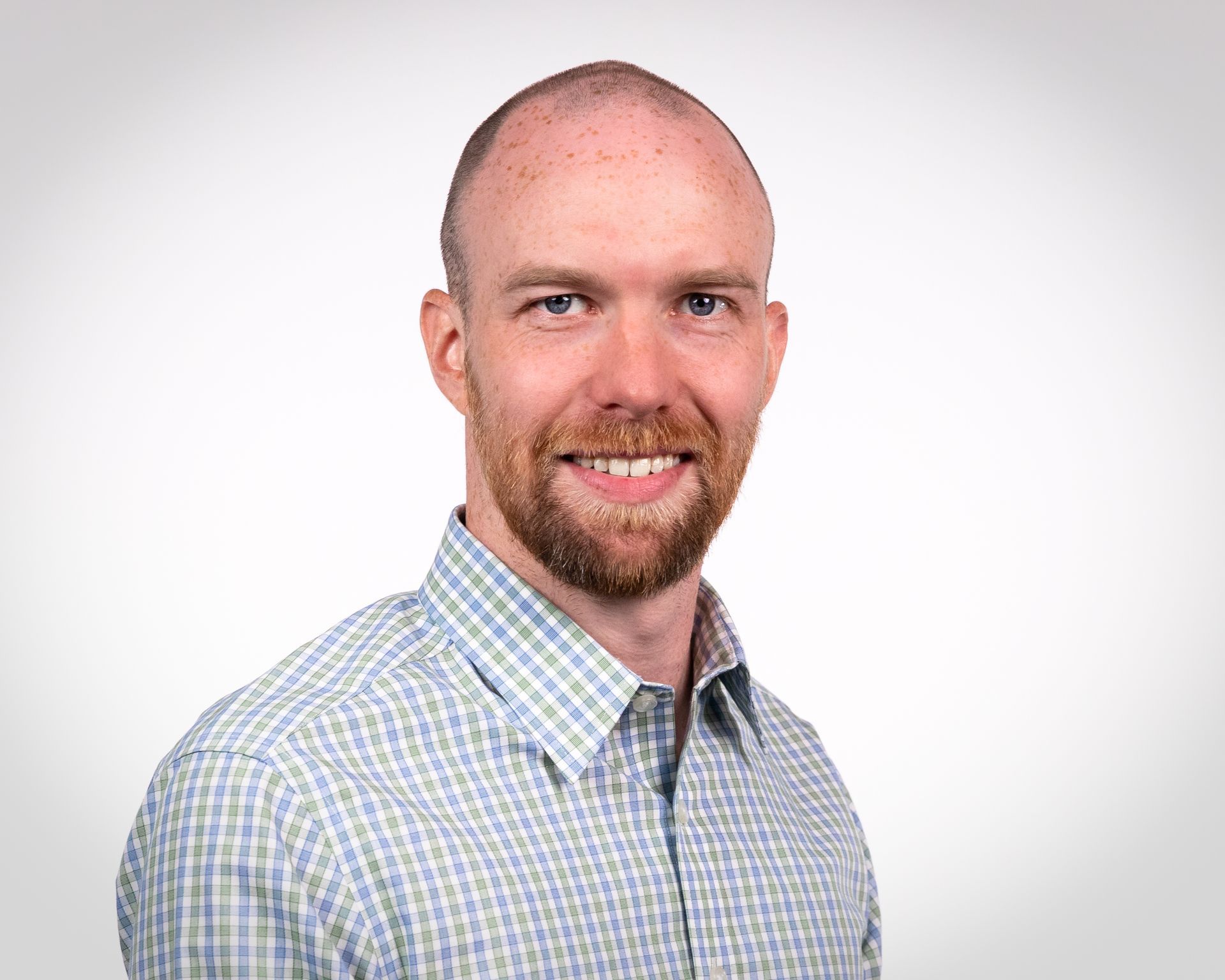 Man with a red beard smiles, wearing a blue and green checkered shirt, against a white background.