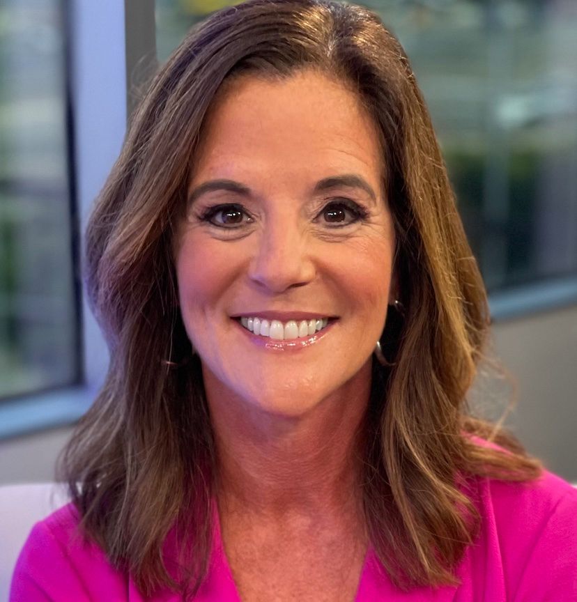 Woman with brown hair smiles, wearing a pink top, lit by studio lighting.