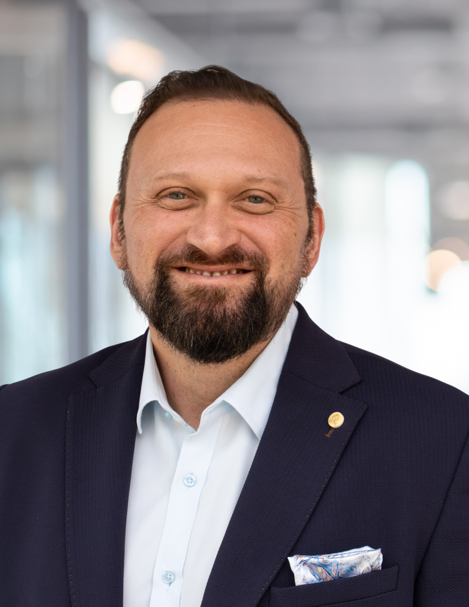 Man in a blue blazer and light blue shirt, smiling with a full beard. Blurred office background.