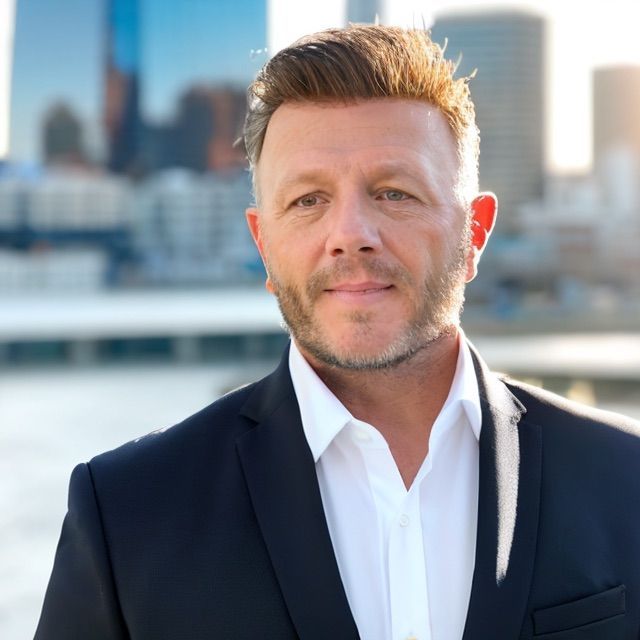 Man in a suit smiles outdoors with a city skyline in the background.