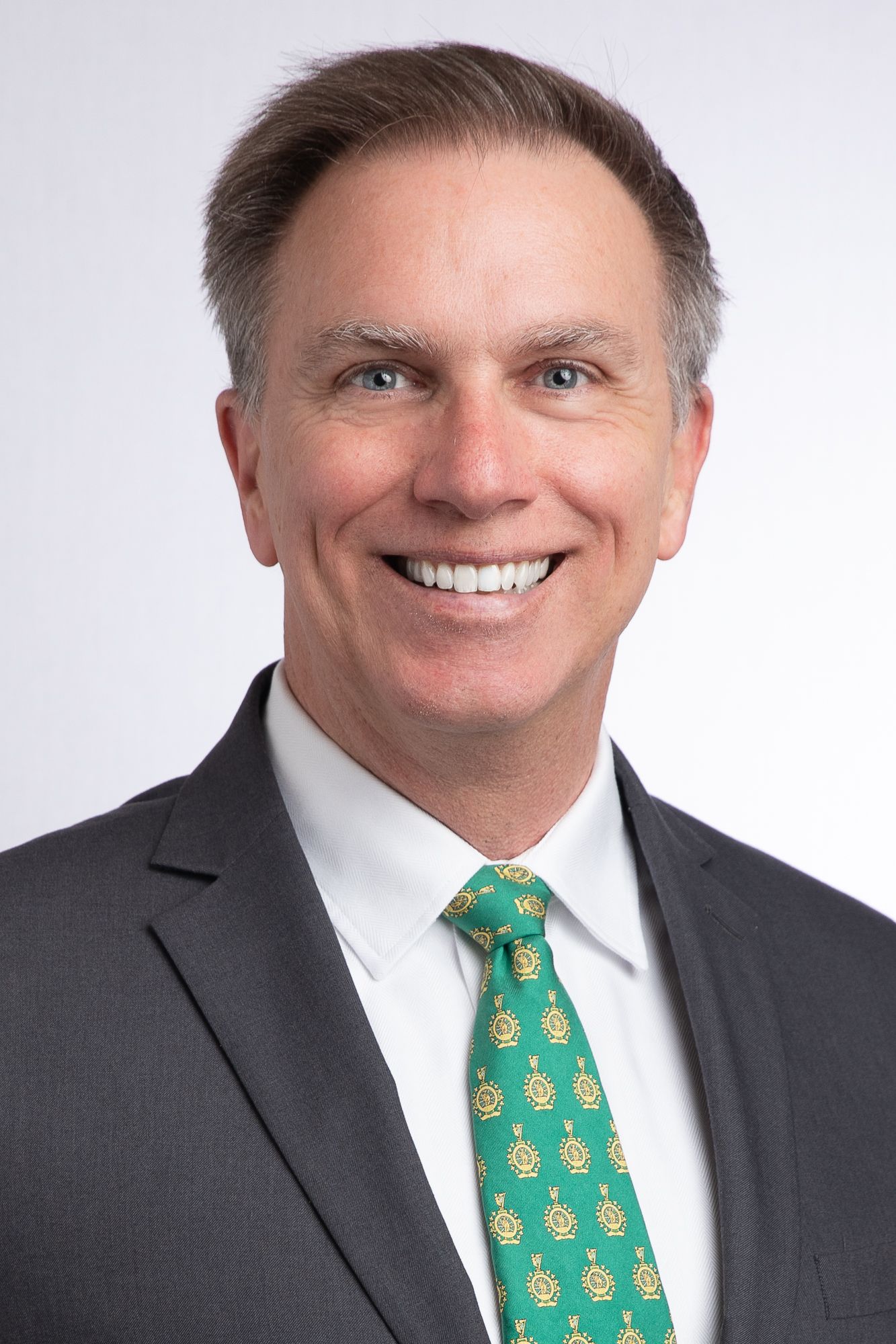 Man in a suit smiles, green tie with gold emblems. Studio background.