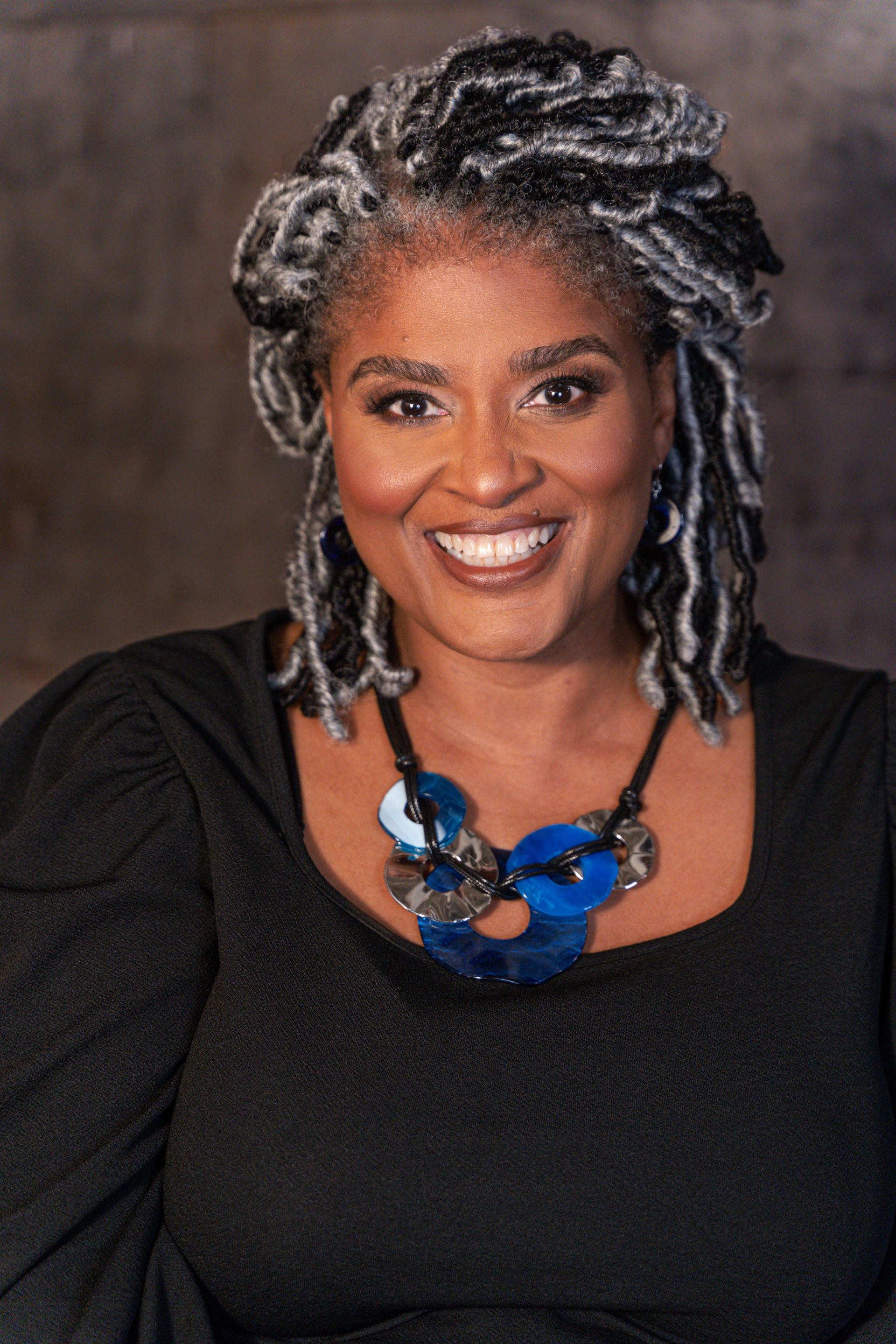 Woman with gray and black locs smiling, wearing a blue necklace and black top.