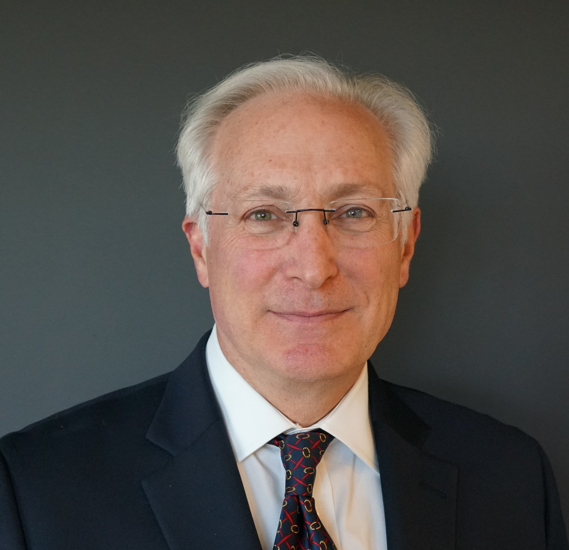 Man in suit and glasses smiles, gray hair, against a gray background.