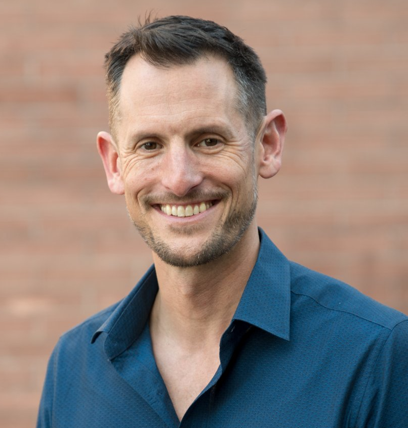 Man in a blue button-down shirt smiles, standing in front of a brick wall.