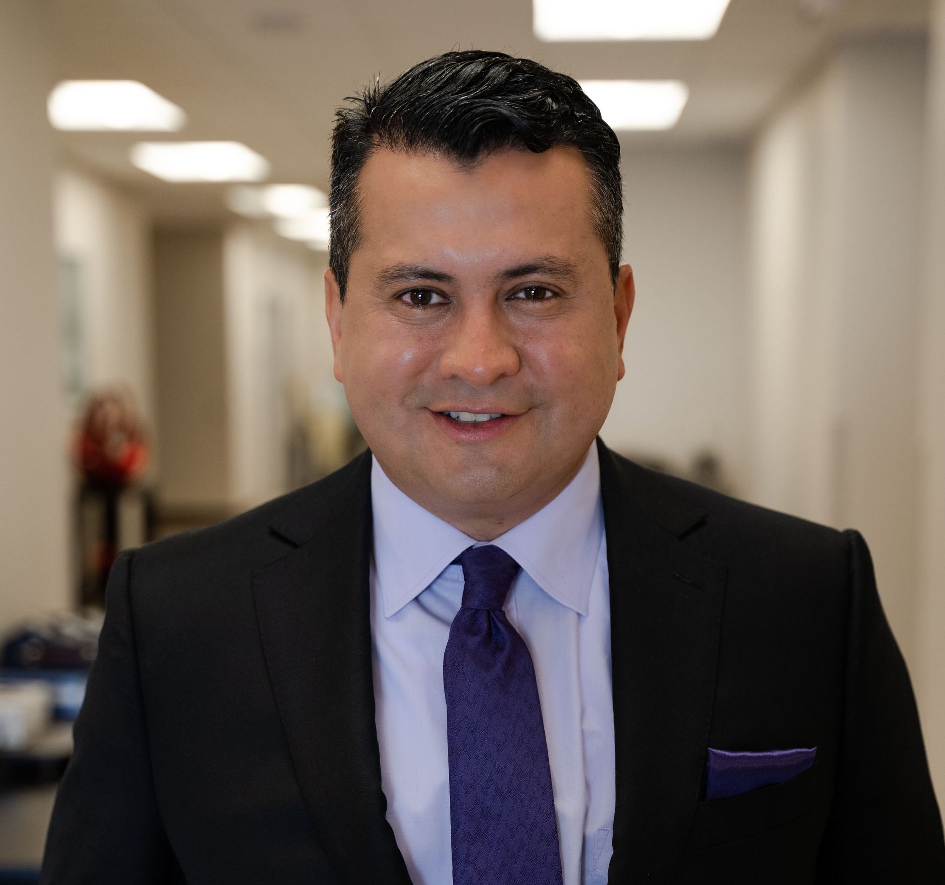 Man in suit smiles, standing in a brightly lit hallway. He wears a purple tie and has a pocket square.