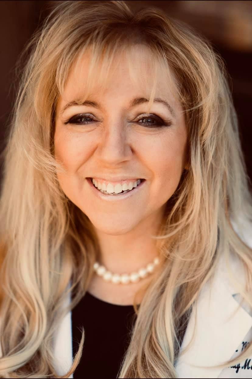 Blonde woman smiling, wearing a pearl necklace, and a white coat.