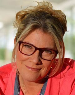 Woman with glasses, blond hair in a messy bun, wearing a coral shirt, smiling.