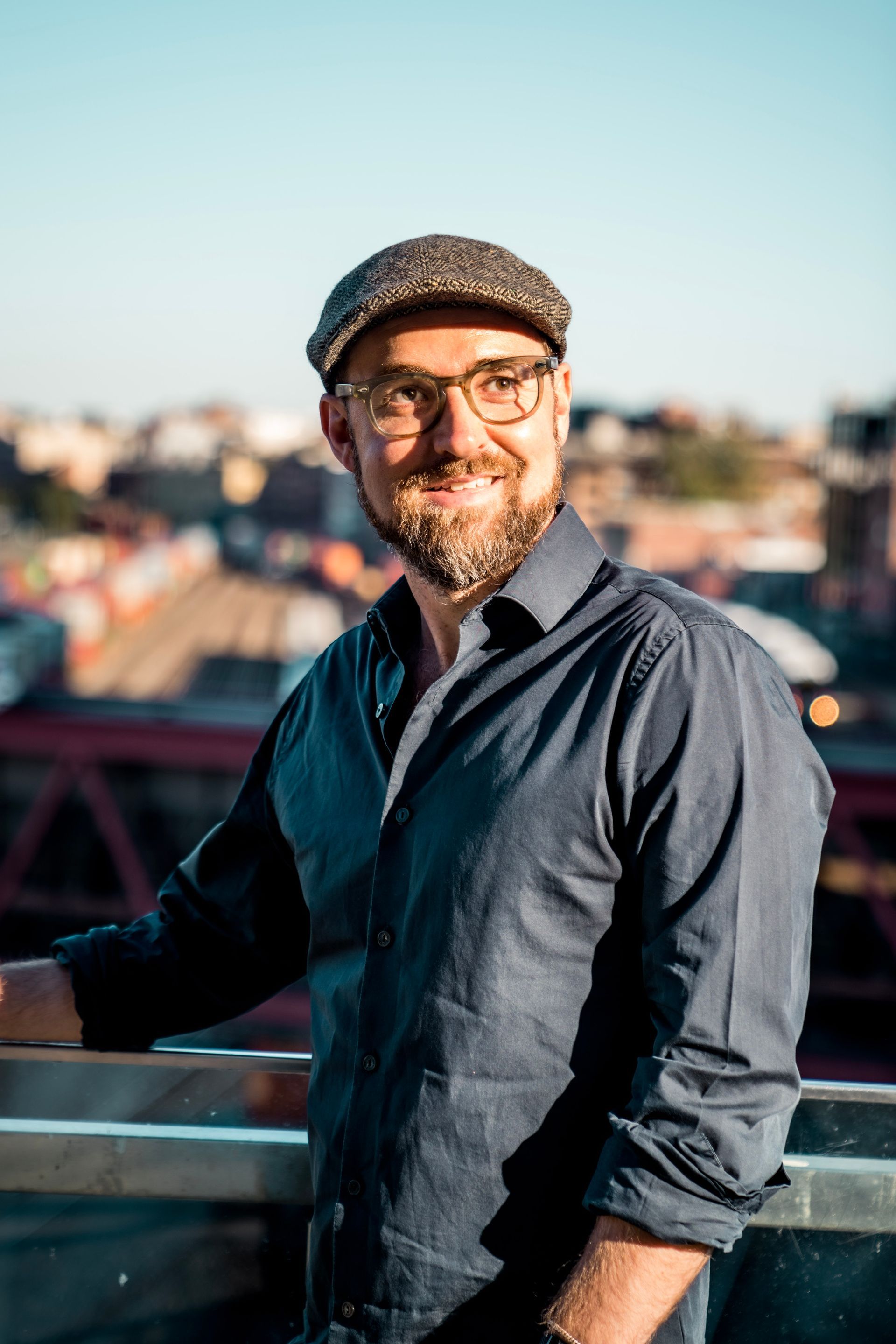 Man with glasses and a cap, smiling, leaning on a railing. City buildings in the background.
