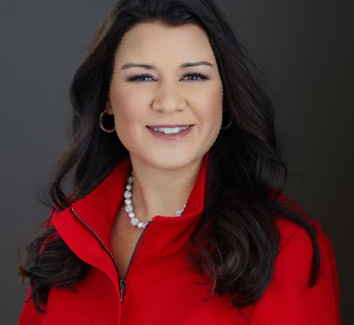 Woman in a red jacket smiles, wearing a pearl necklace, hoop earrings, and has dark hair; studio shot.