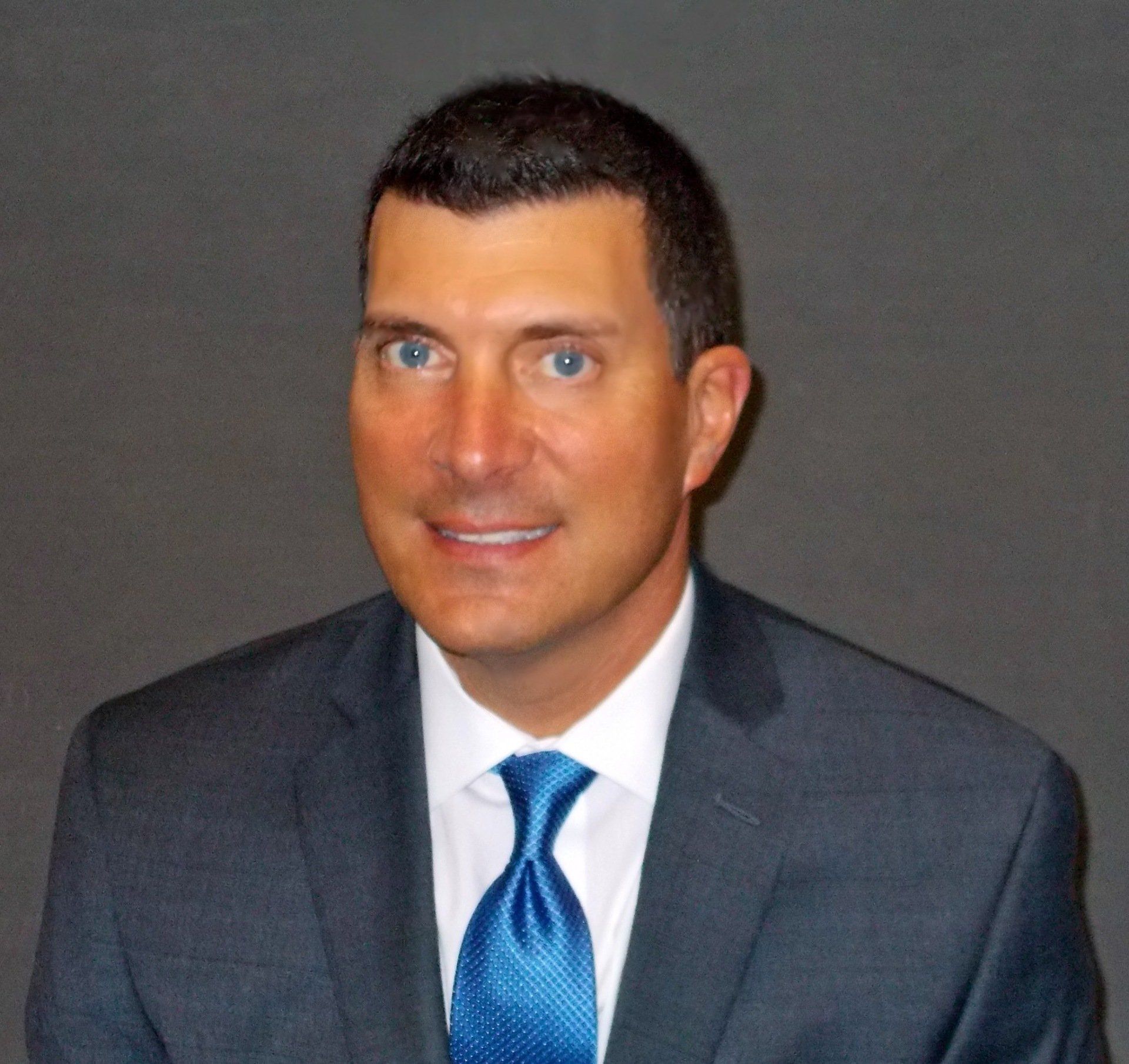 Man in a gray suit and blue tie smiles, against a gray background.