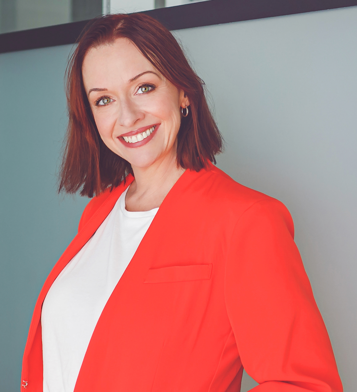 Woman with red hair, wearing a red blazer over a white shirt, smiles at the camera.