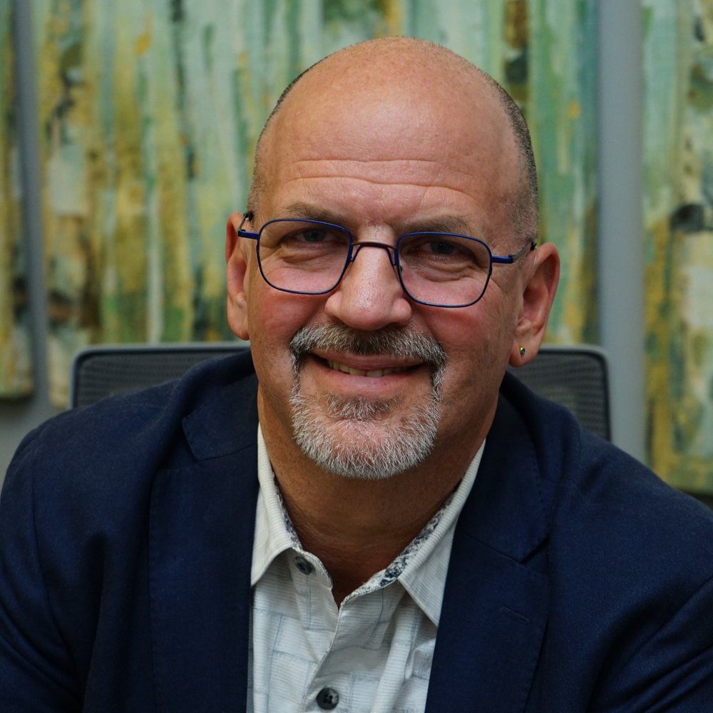 Man in glasses smiles, wearing a blue blazer and patterned shirt, indoors in front of abstract art.