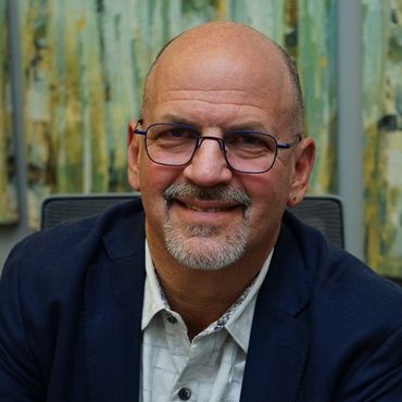 Man in glasses smiles, wearing a blue blazer and patterned shirt, indoors in front of abstract art.