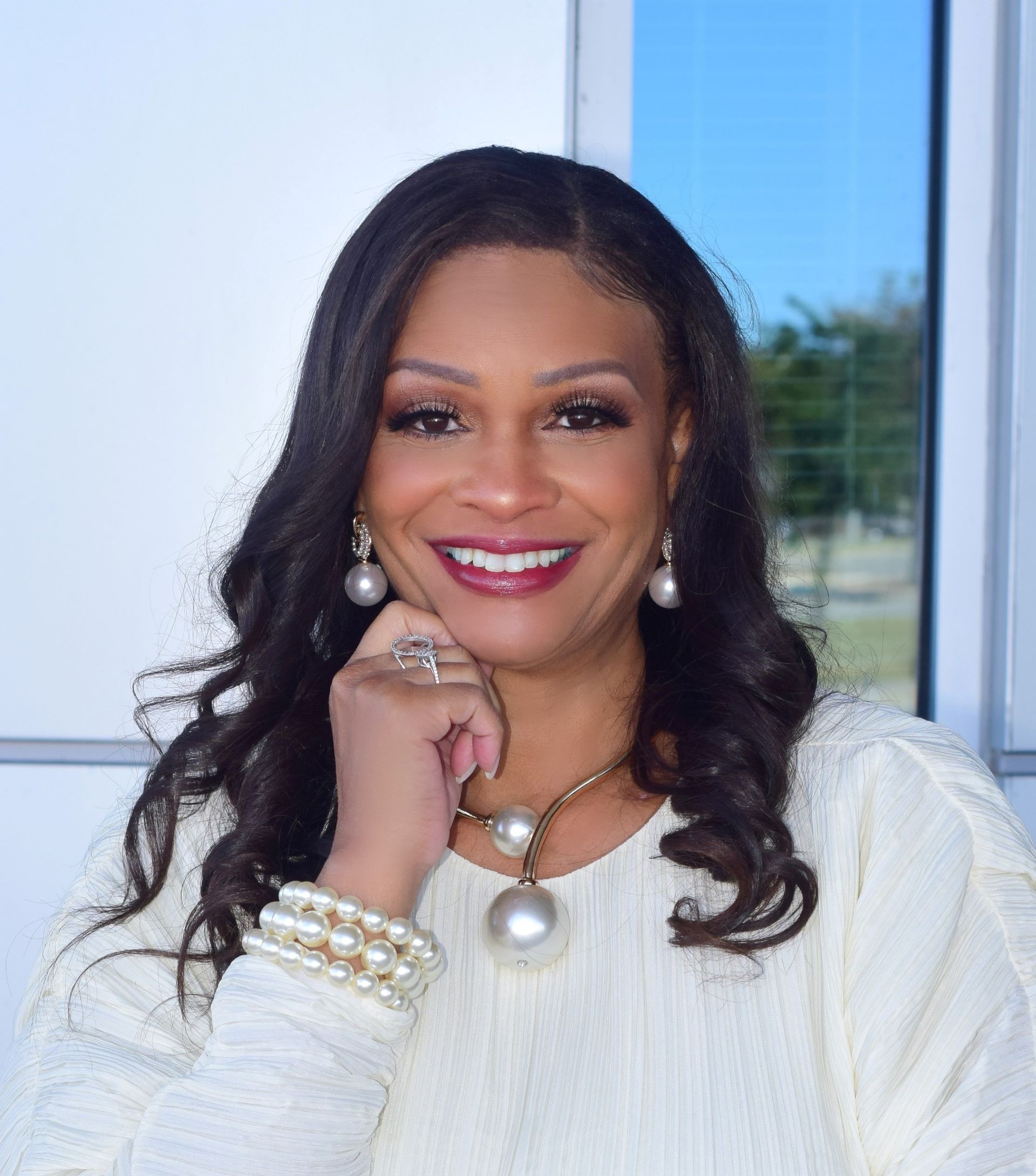 Woman in white outfit smiles, resting hand on face, wearing pearl jewelry, in a bright setting.