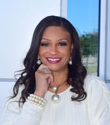 Woman in white outfit smiles, resting hand on face, wearing pearl jewelry, in a bright setting.