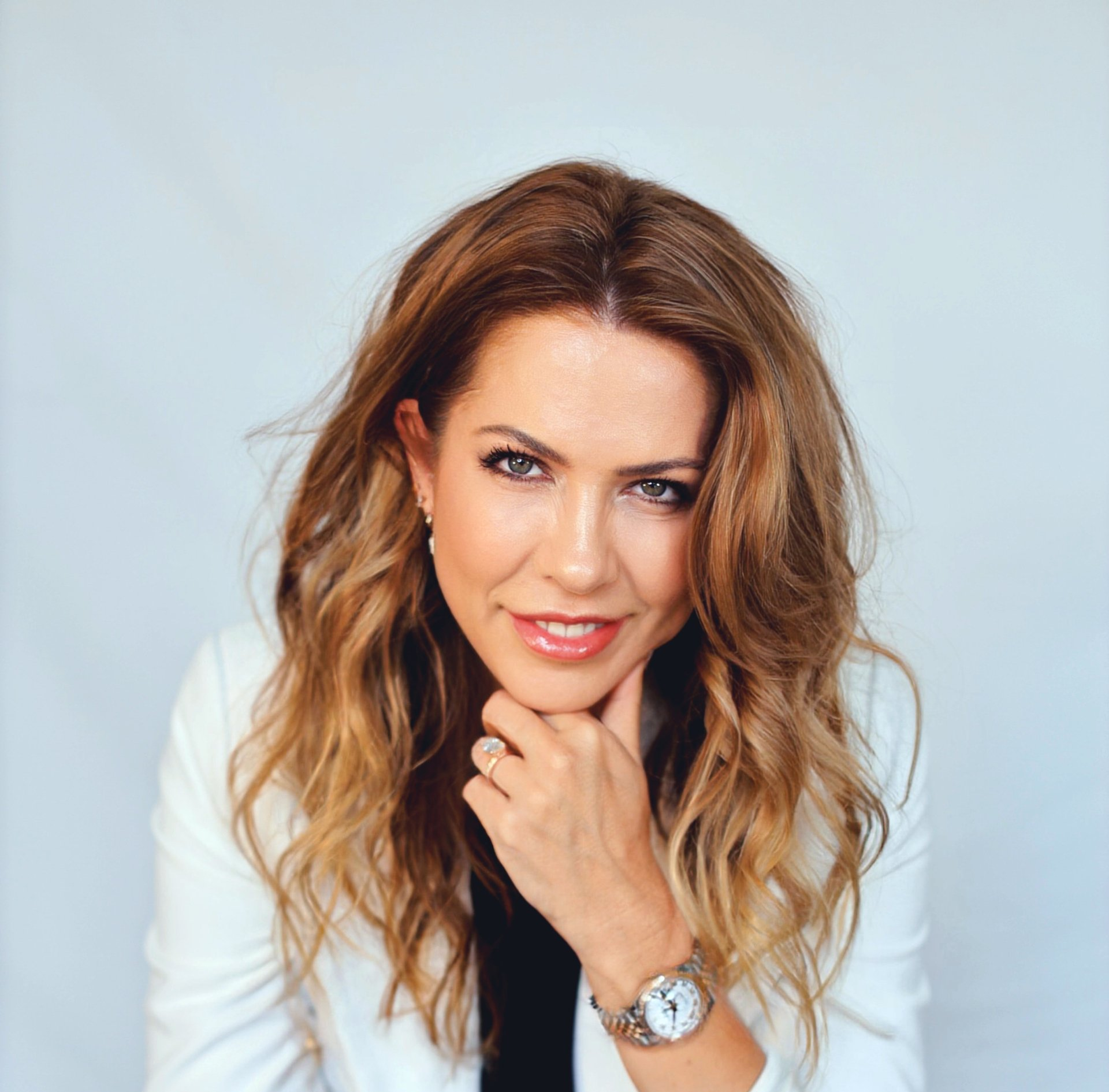 Woman with wavy brown hair, wearing a white blazer, resting chin on hand, smiling.